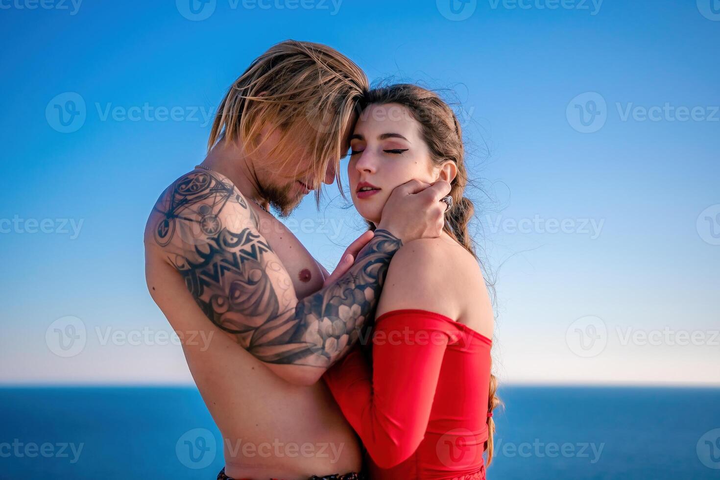Portrait of tattooed man and sensual young brunette woman in red dress, happily dancing outdoors isolated on blurry sea background with natural bokeh in soft warm sunset backlight. Selective focus. photo
