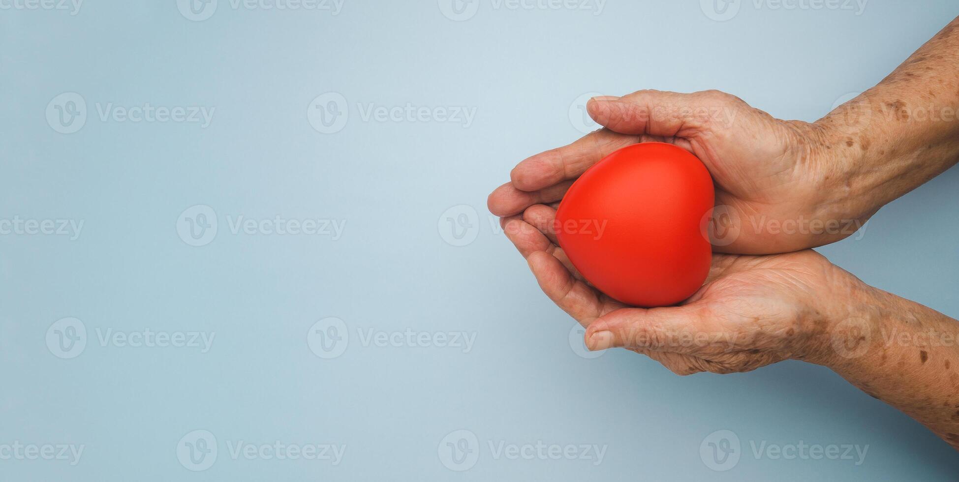 A red heart on the palm of a senior woman is isolated on light blue background. Healthcare, wellbeing, World heart day, World health day. Valentine's day. People, age, love, and health care concept photo