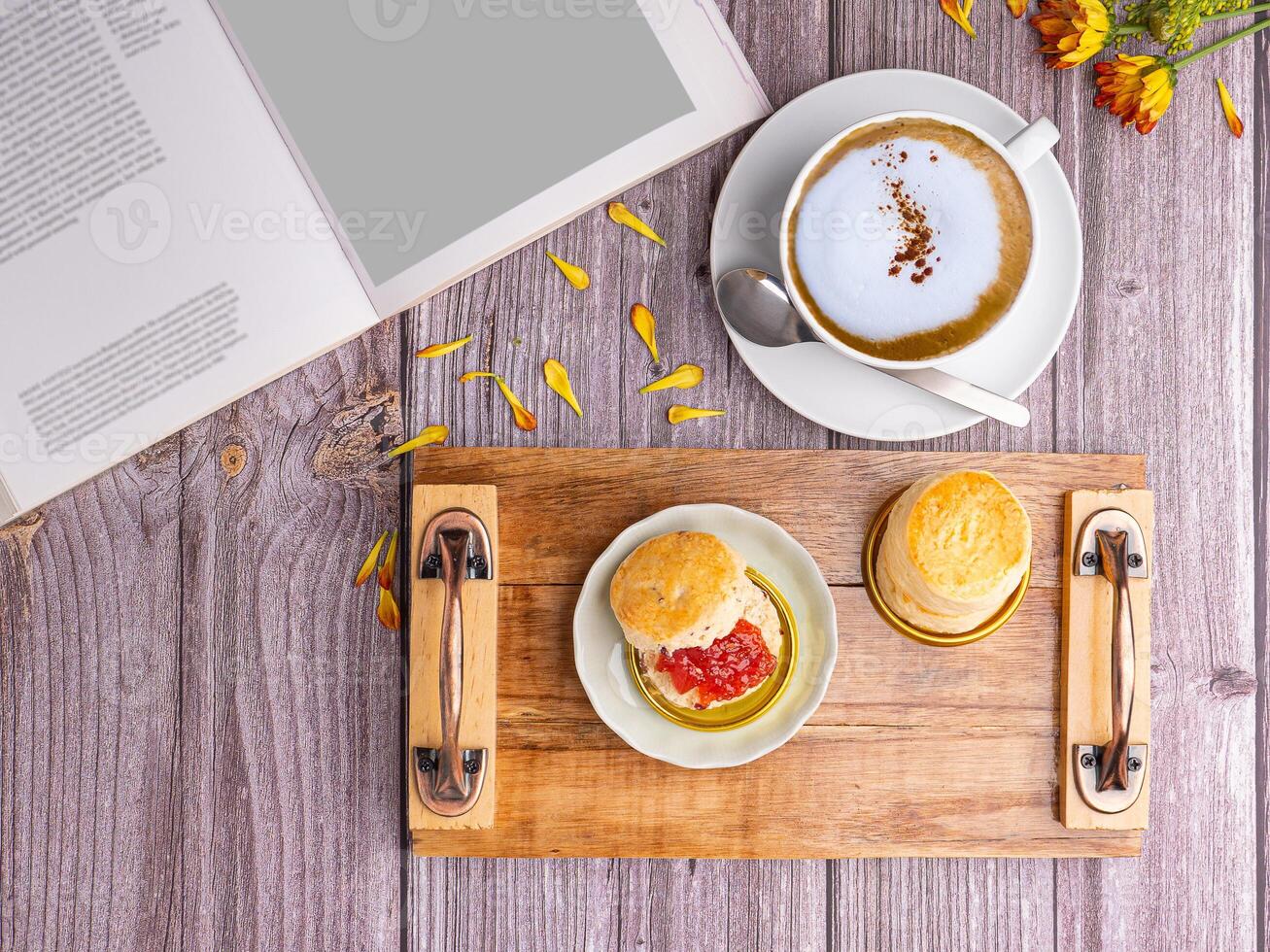 Scones traditional English delicious freshly baked homemade with strawberry jam served on a wooden tray with a coffee cup and a book on a wooden table. Top view. Relaxation concept photo