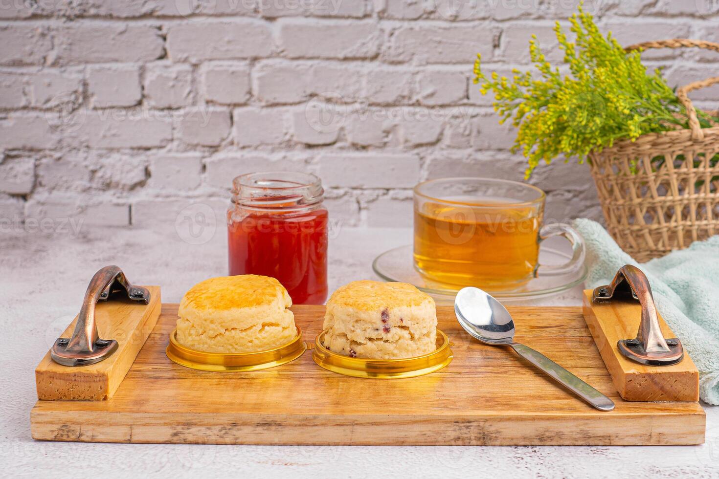 English style scones delicious freshly baked homemade, strawberry jam placed on a wooden tray with a cloth and a teacup with a white brick wall. Side view. Dessert, beverage, and relaxation concept photo