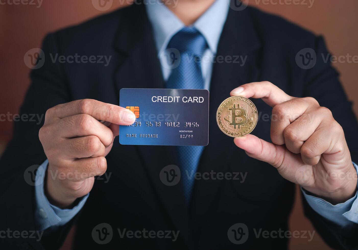 Hand of a businessman holding a bitcoin golden coin and a mockup blue credit card while standing with a brown background. Close-up photo. Virtual cryptocurrency and future money concept photo