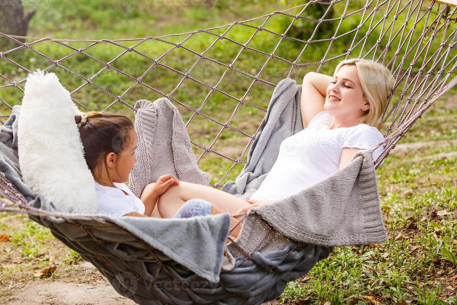 Wide view of a young mother and daughter relaxing together and smiling sitting in a hammock, hugging and lounging during a sunny summer day in a holiday home garden with grass and trees, lifestyle photo