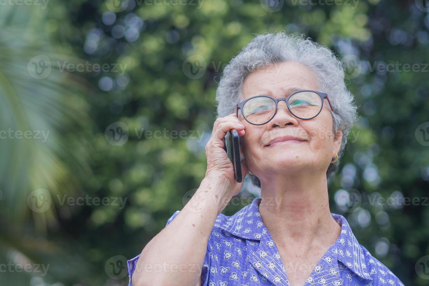 Happy beautiful senior woman is using a smartphone and looking up while standing in a garden. Space for text. Aged people and communication concept photo