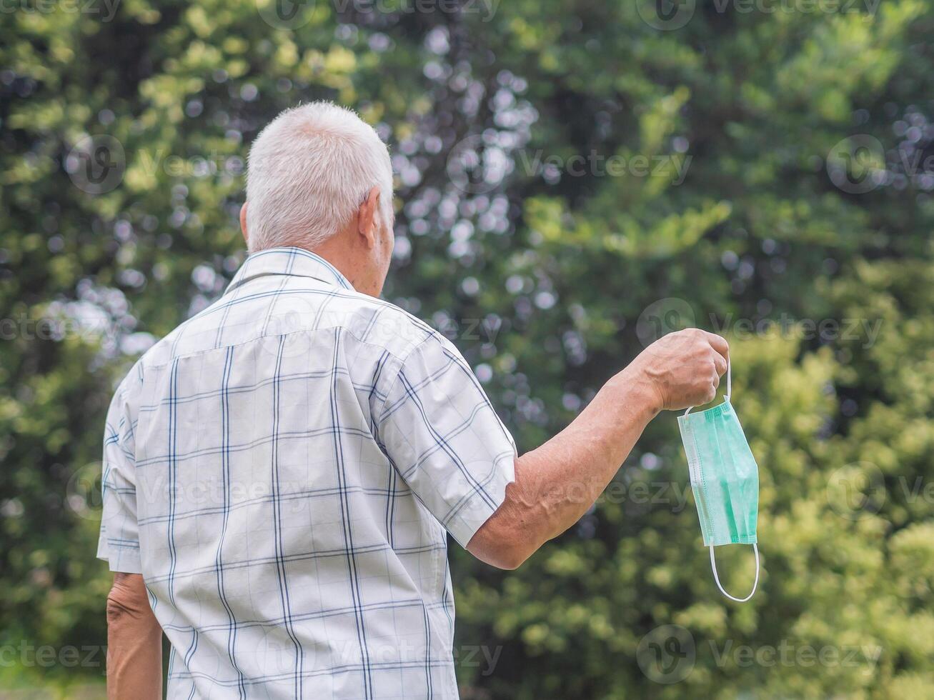 Back view of a senior man with short gray hair holding a face mask while standing outdoor. Space for text. Aged people and healthcare concept photo