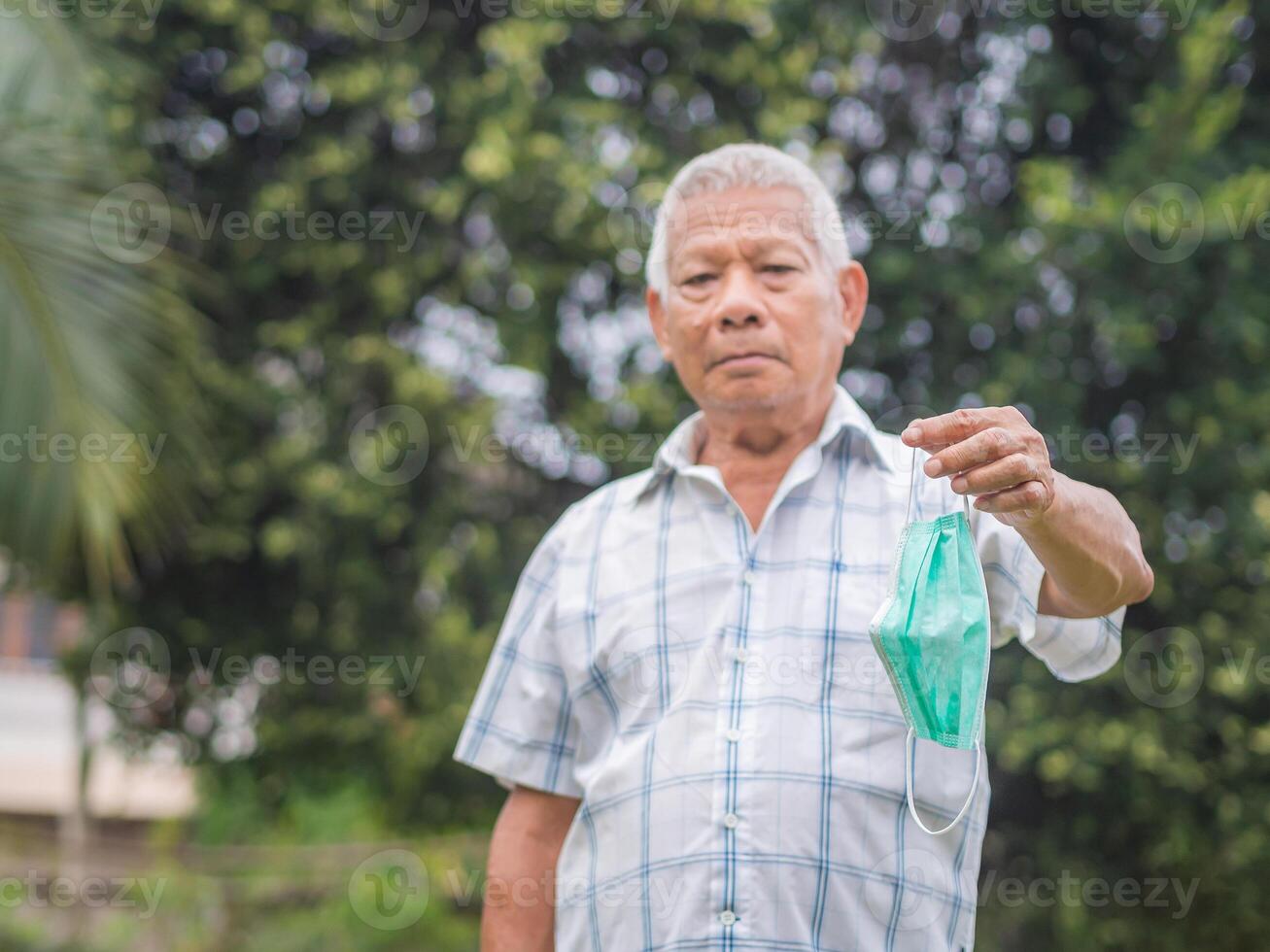 Portrait of a senior man holding a face mask while standing outdoor. Space for text. Selective focus with blurred background. Aged people and healthcare concept photo