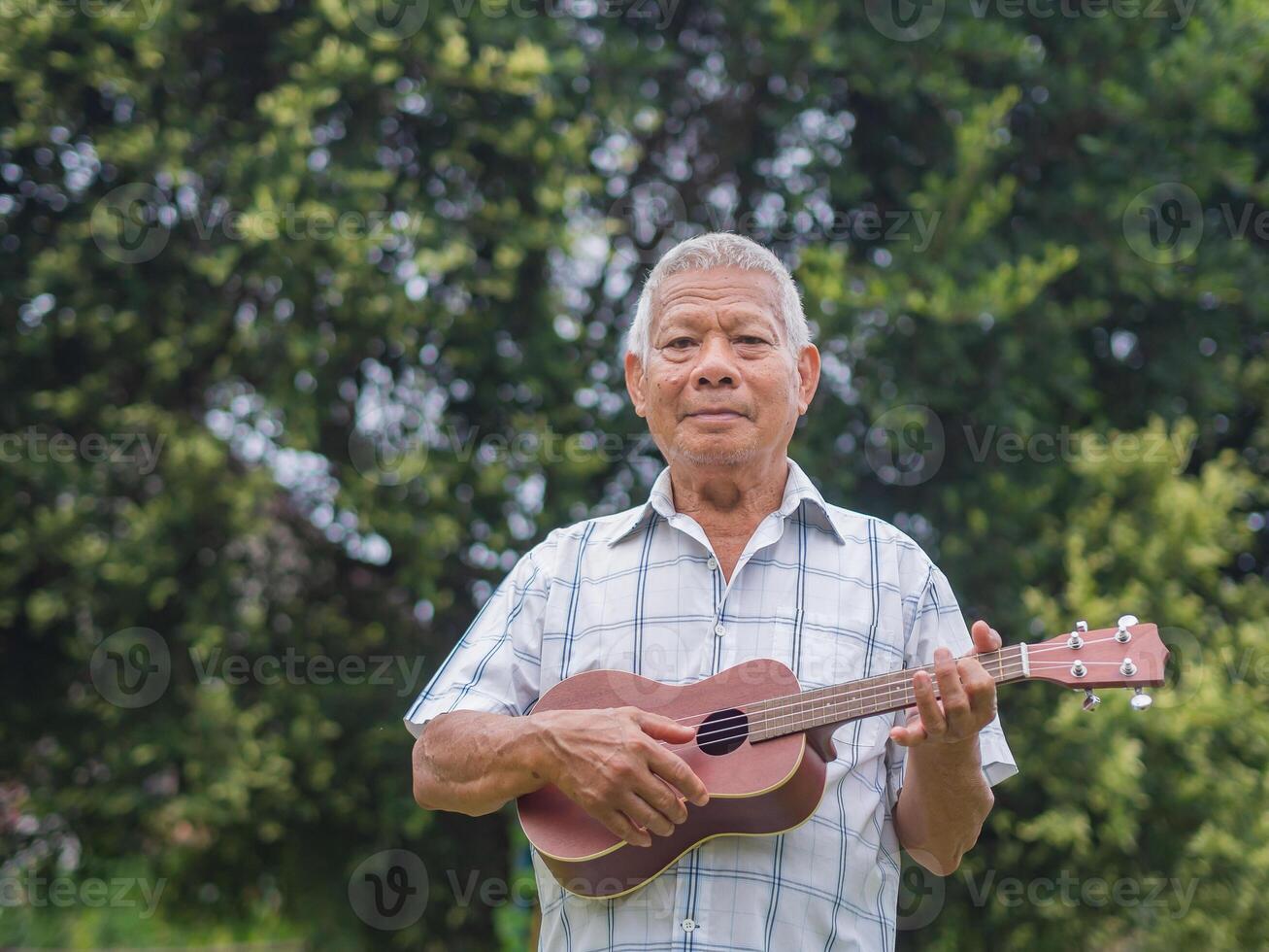 Happy elderly man with short gray hair playing the ukulele, smiling and looking at the camera while standing in a garden. Enjoy life after retiring. Concept of aged people and relaxation photo