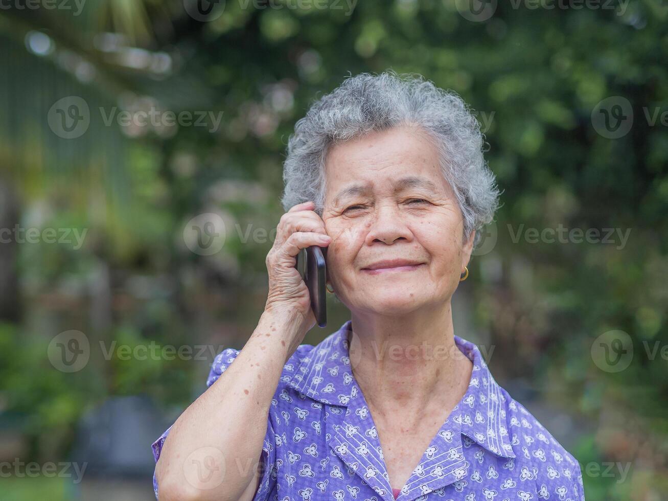 Happy beautiful senior woman is using a smartphone, smiling and looking at the camera while standing in a garden. Space for text. Aged people and communication concept photo