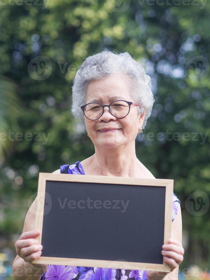 Portrait of an elderly Asian woman with short gray hair holding a mini blackboard, smiling and looking at the camera while standing in a garden. Aged people and relaxation concept photo