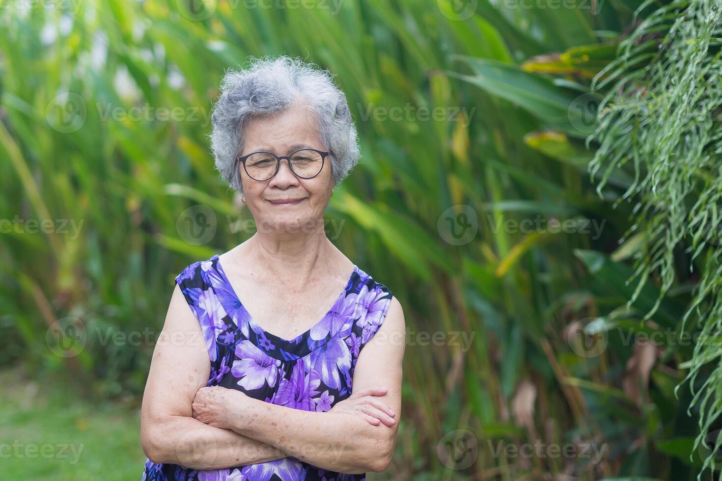 Portrait of a senior woman with short gray hair, wearing glasses, smiling and looking at the camera while standing in a garden. Aged people and relaxation concept photo