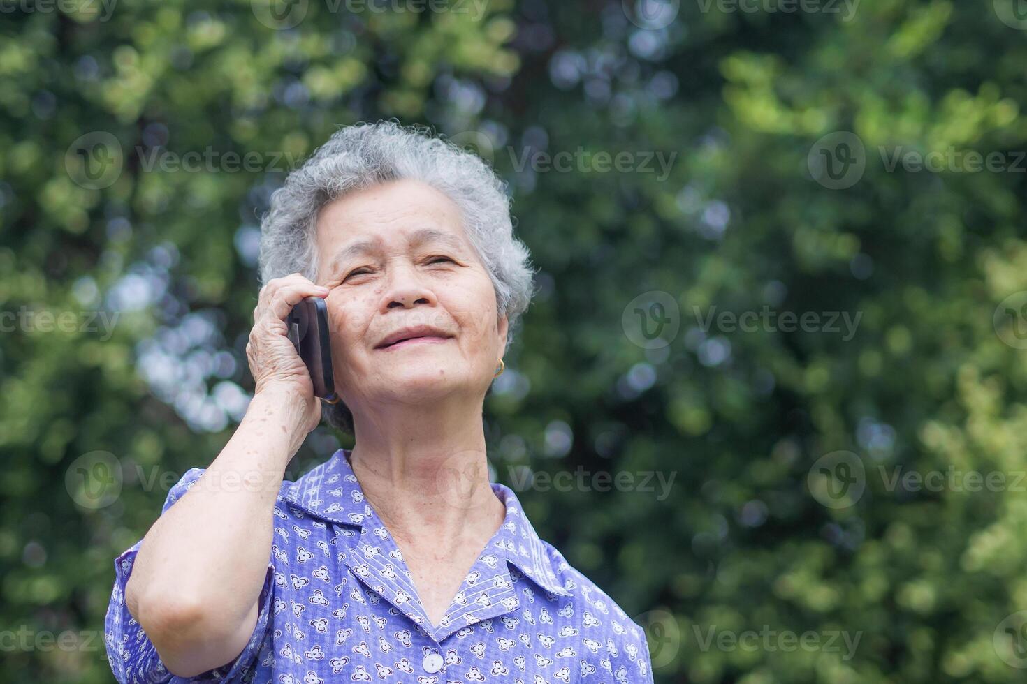Happy beautiful senior woman is using a smartphone and looking up while standing in a garden. Space for text. Aged people and communication concept photo