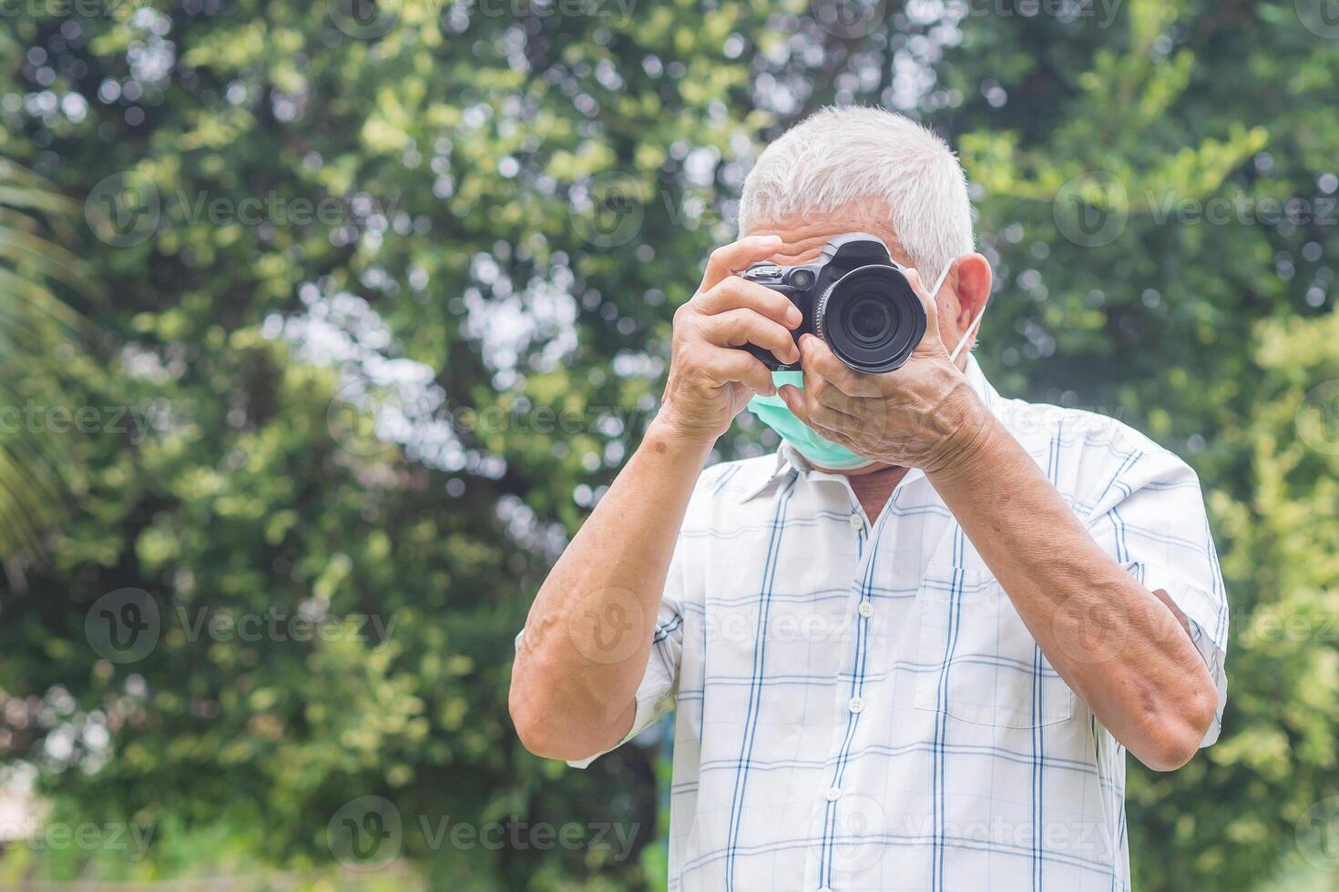 Senior man wearing a face mask and taking photographs with a digital camera while standing outdoors. Aged people photographer and relaxation concept photo