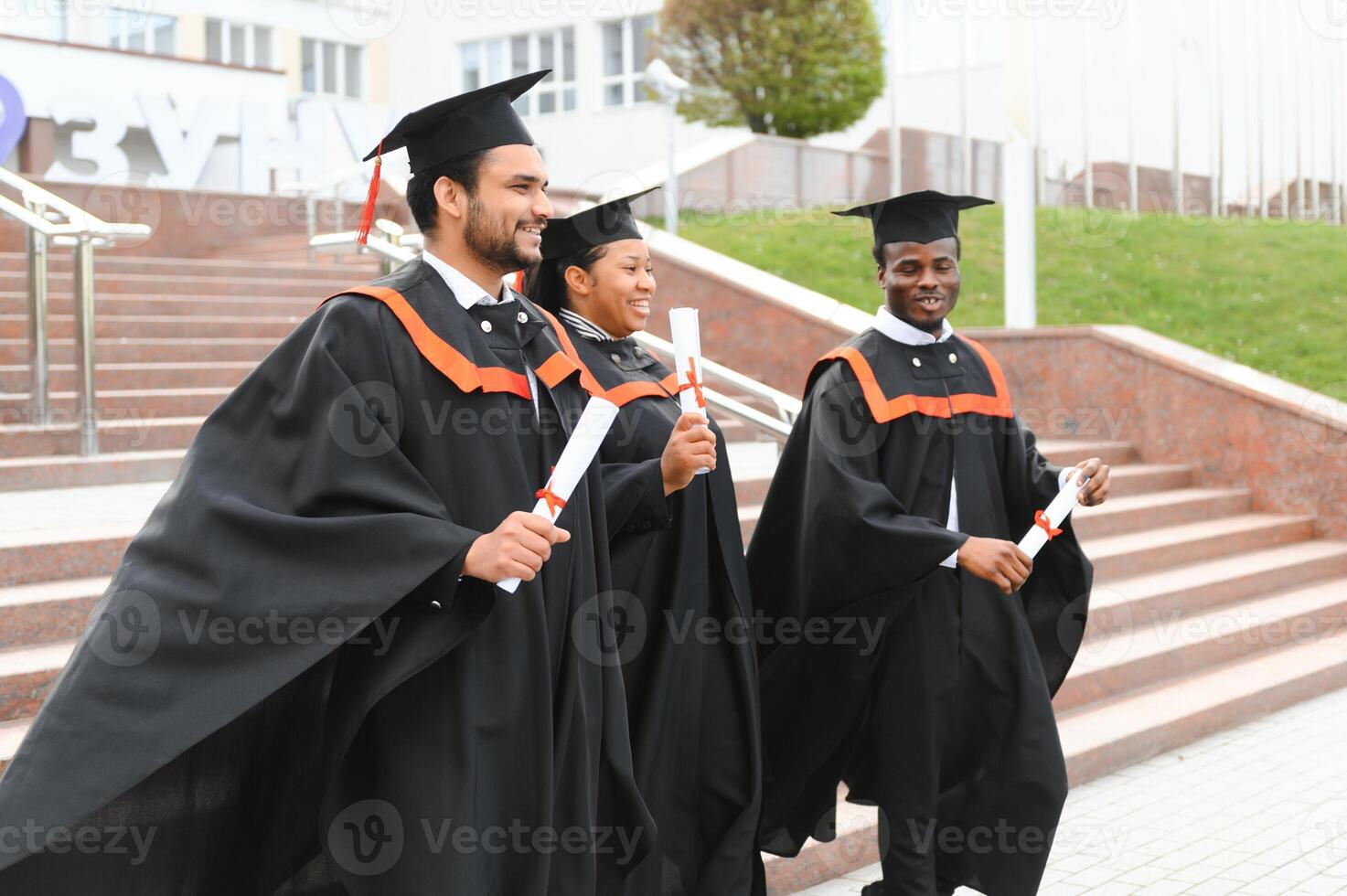 Group of happy smiling multicultural people in graduation gowns and caps with diplomas in hands graduate university outdoors in campus looking at camera. Graduation from college, university concept. photo