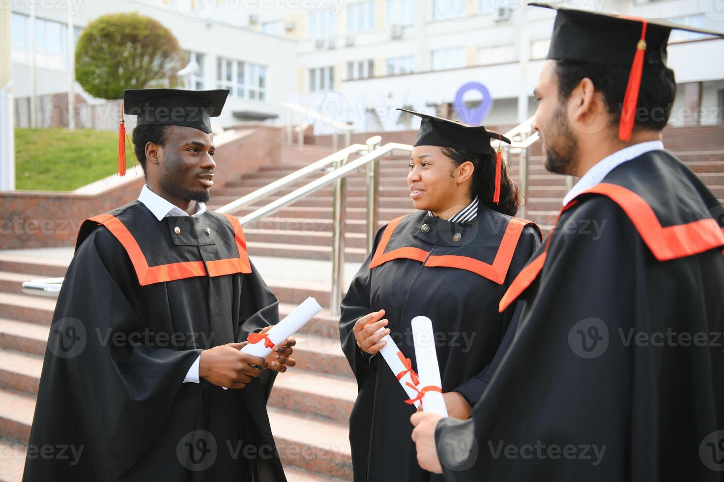 Group of happy smiling multicultural people in graduation gowns and caps with diplomas in hands graduate university outdoors in campus looking at camera. Graduation from college, university concept. photo