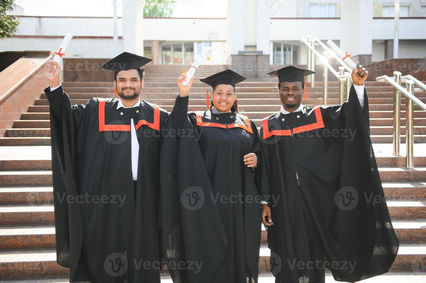 Group of happy smiling multicultural people in graduation gowns and caps with diplomas in hands graduate university outdoors in campus looking at camera. Graduation from college, university concept. photo