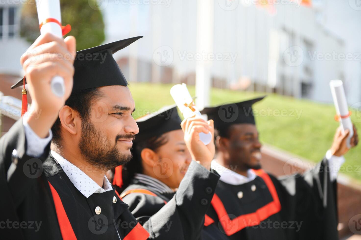 Group of happy smiling multicultural people in graduation gowns and caps with diplomas in hands graduate university outdoors in campus looking at camera. Graduation from college, university concept. photo