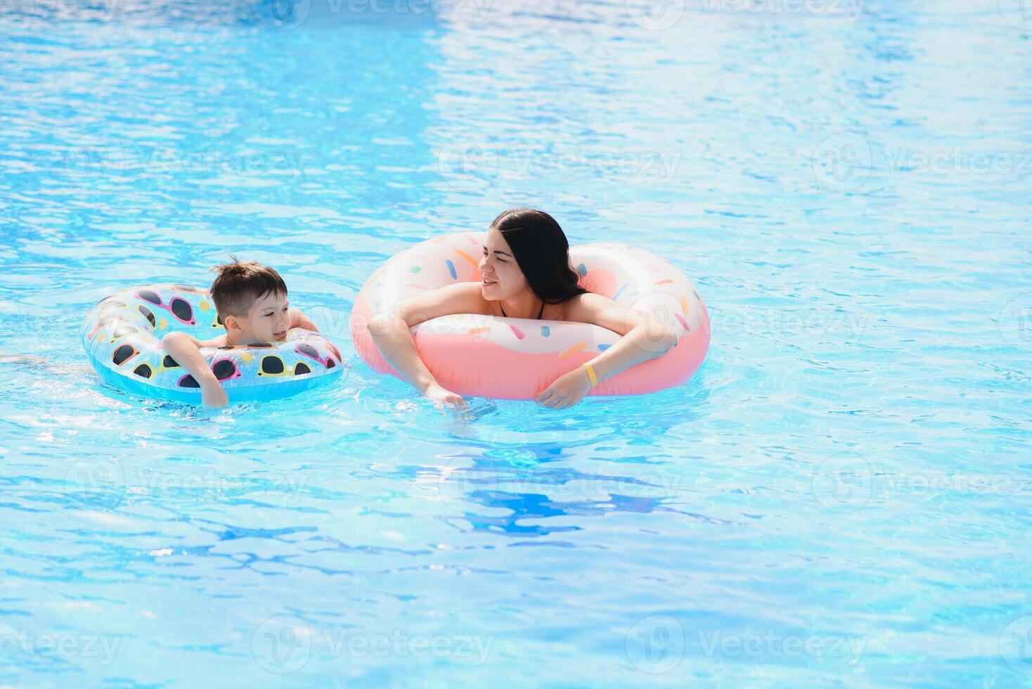 Happy family, young active mother and adorable curly little baby having fun in a swimming pool, child learning to swim in an inflatable toy ring, enjoying summer vacation at a tropical resort photo