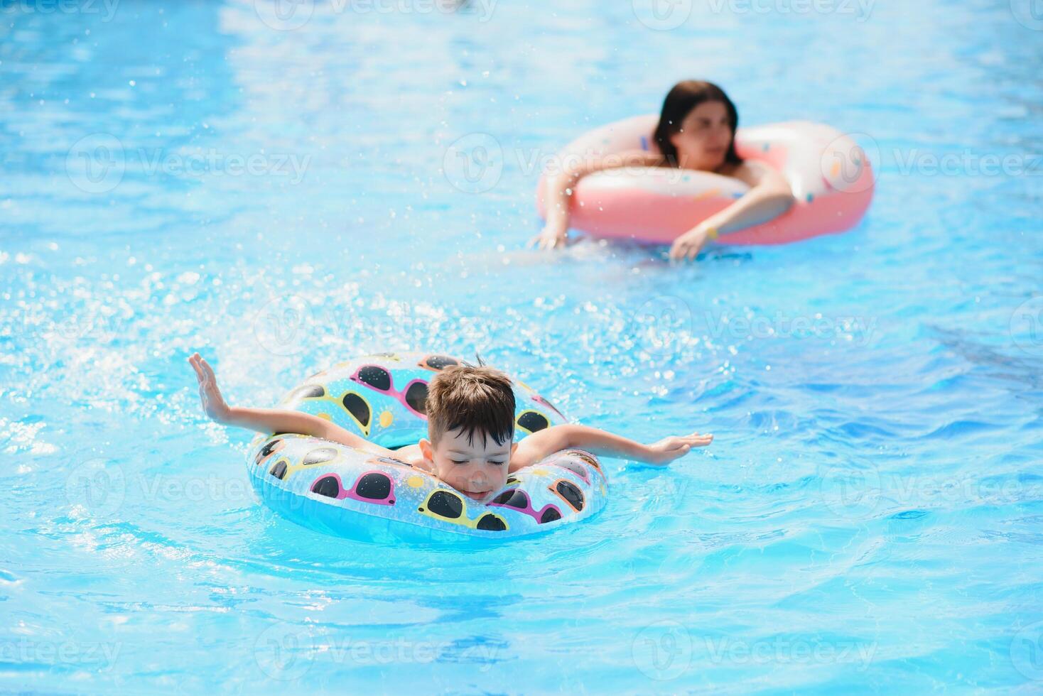 Happy family, young active mother and adorable curly little baby having fun in a swimming pool, child learning to swim in an inflatable toy ring, enjoying summer vacation at a tropical resort photo