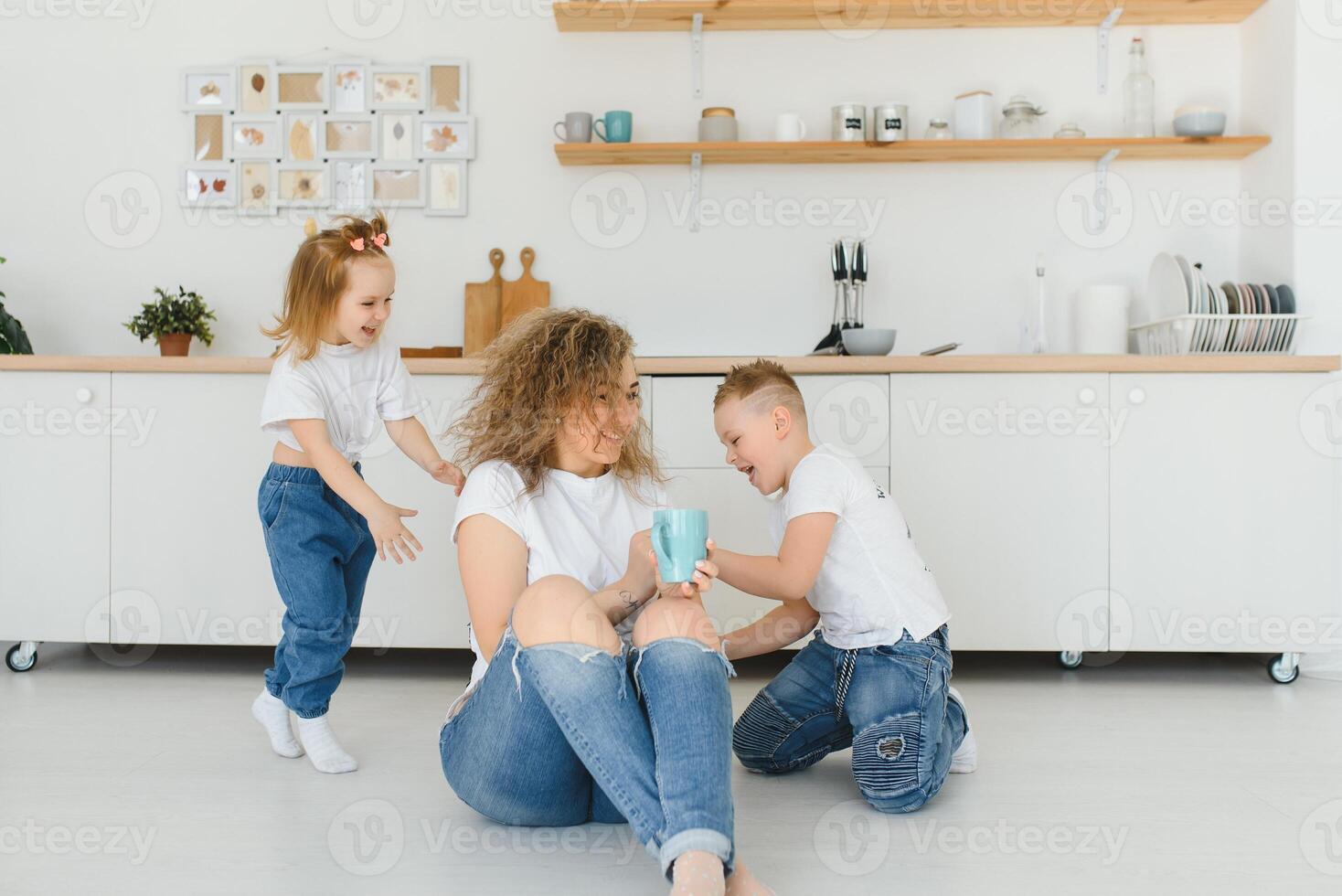 Happy mother hugging daughter and son sitting on wooden floor in modern kitchen at home. Mom hugging children. Happy family relationship concept photo