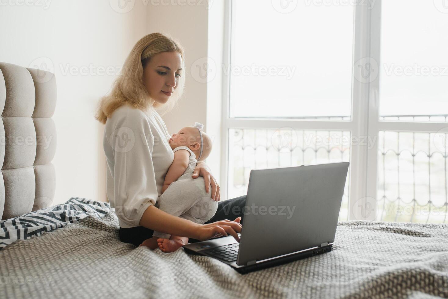 white young mother sitting with baby on bed and working at laptop, work at home photo