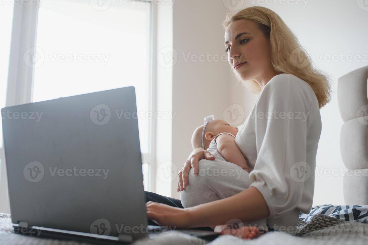 white young mother sitting with baby on bed and working at laptop, work at home photo
