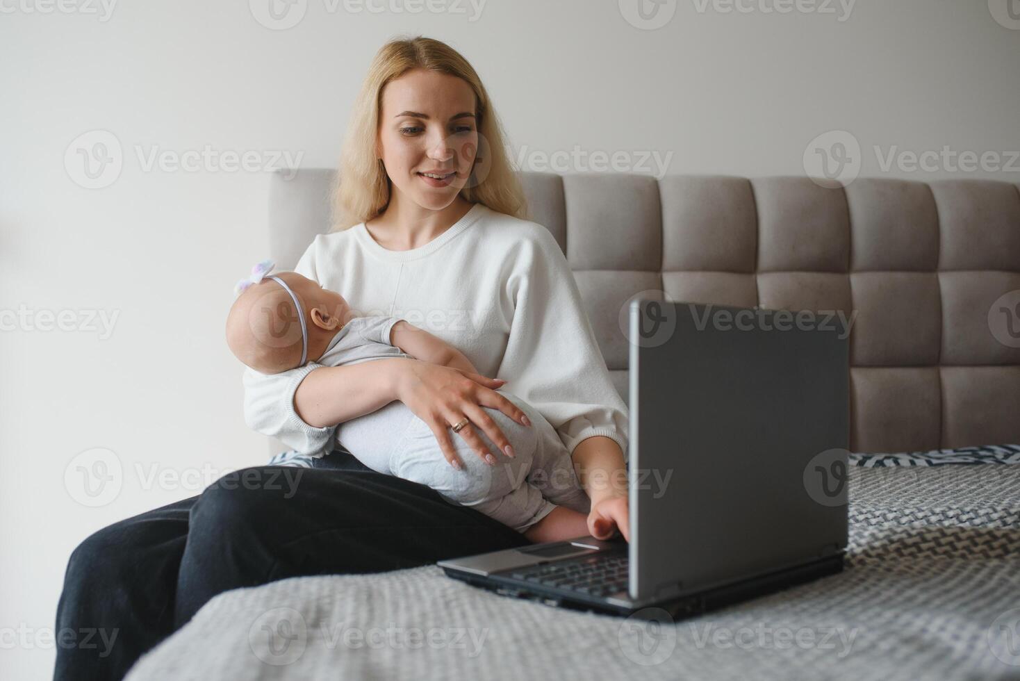 white young mother sitting with baby on bed and working at laptop, work at home photo