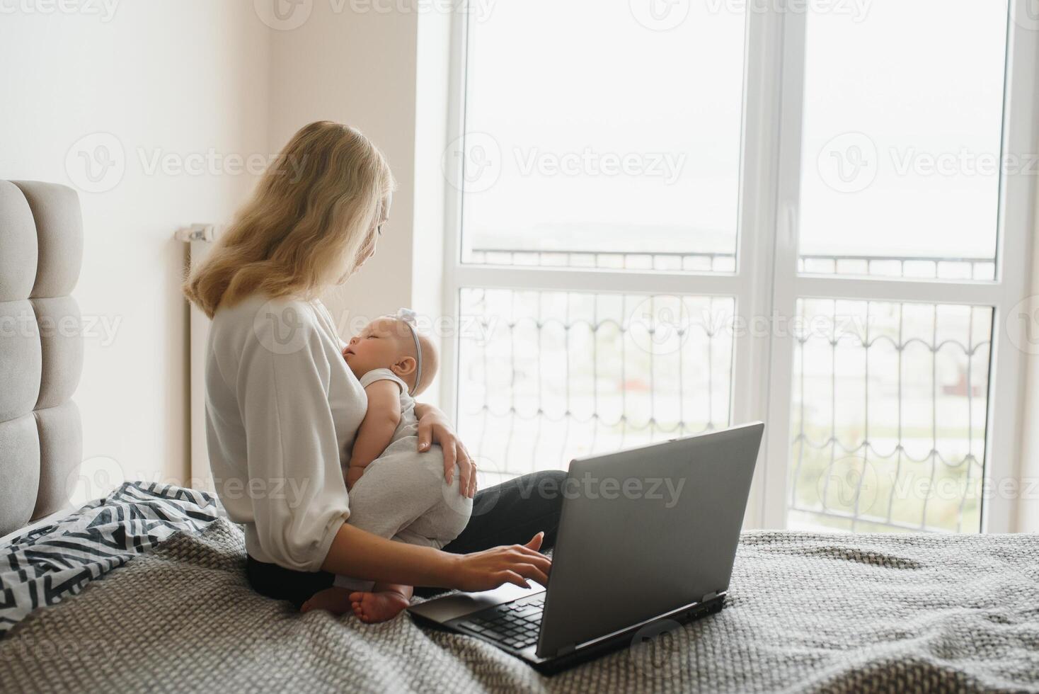 white young mother sitting with baby on bed and working at laptop, work at home photo