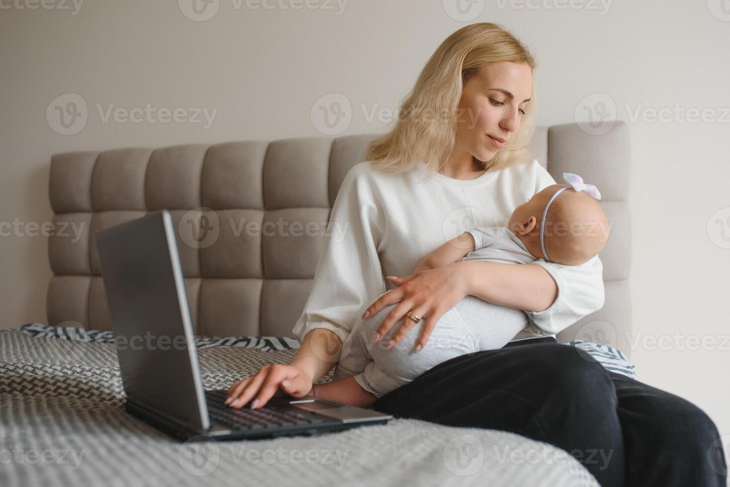 white young mother sitting with baby on bed and working at laptop, work at home photo