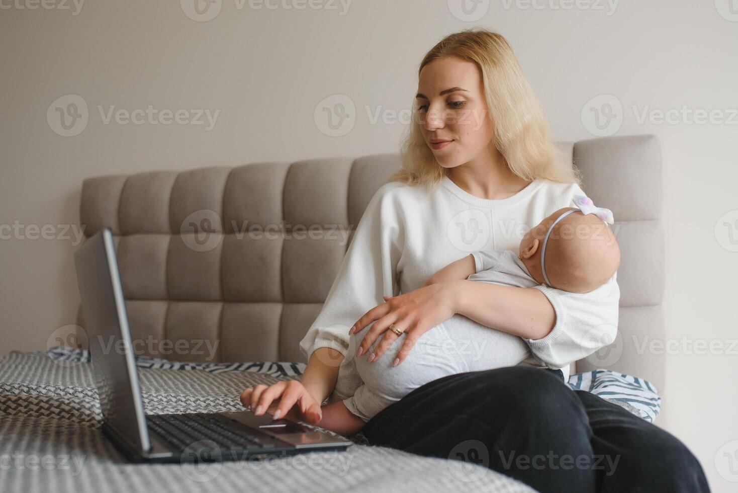 white young mother sitting with baby on bed and working at laptop, work at home photo