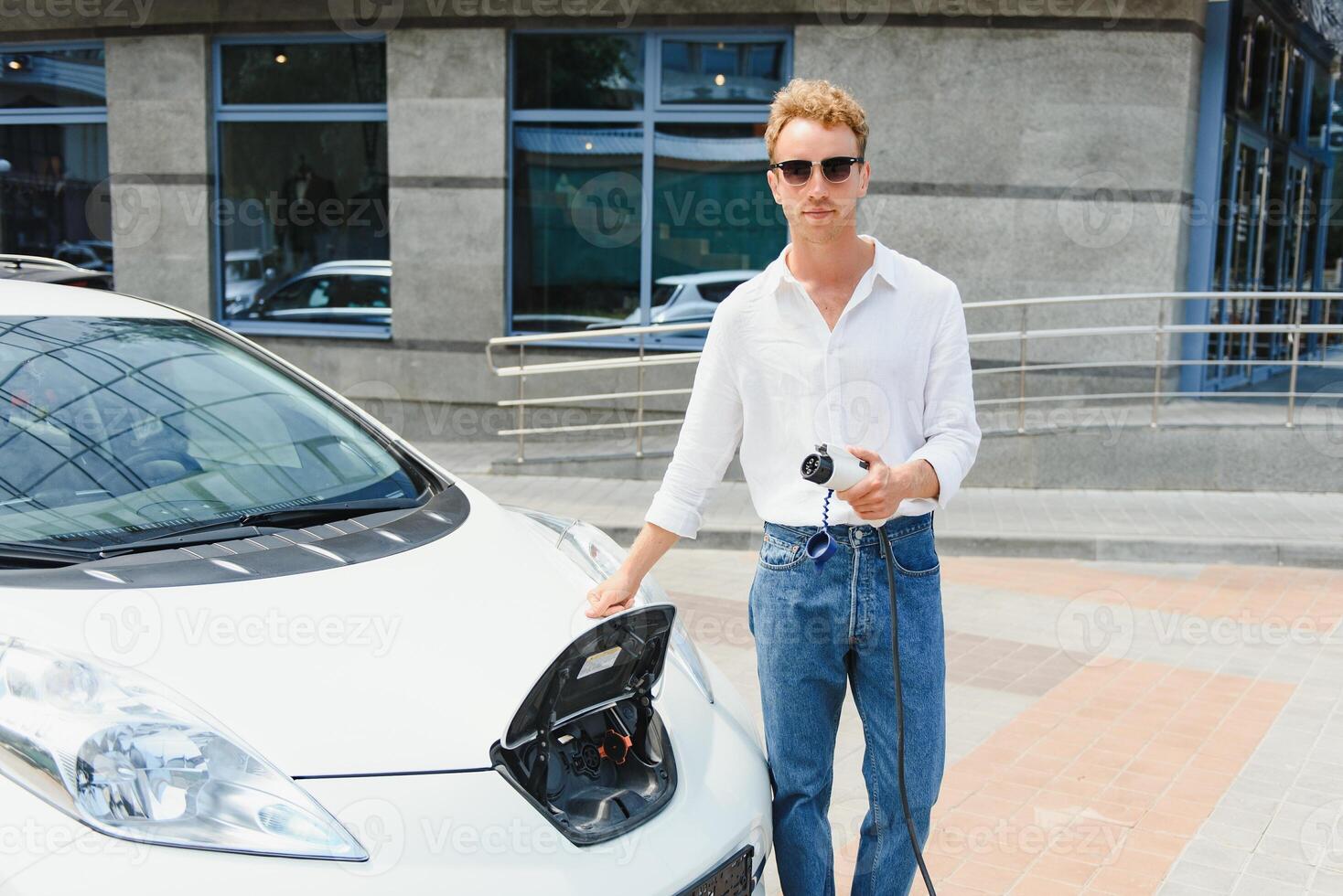 Stylish man with coffe cup in hand inserts plug into the electric car charging socket photo