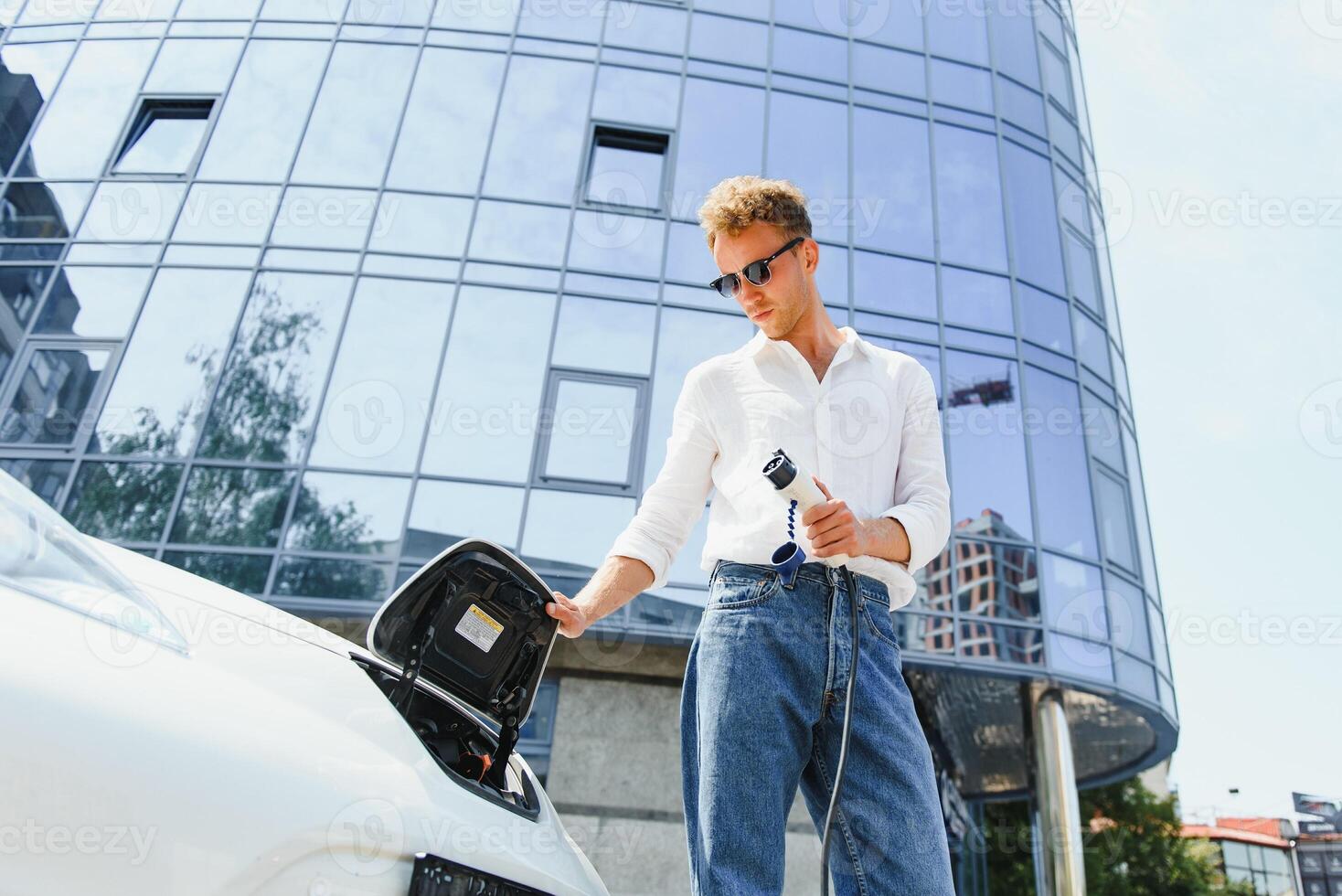 young handsome man holding charging cable at electric charging station point standing near his new car , looking satisfied. photo