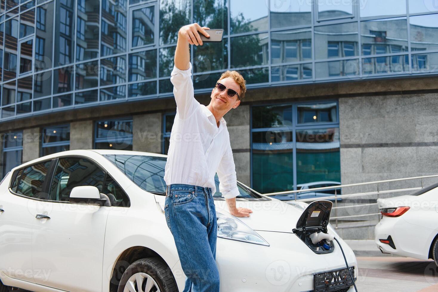 Portrait of young handsome european man, making selfie photo on his smartphone while leaning on his electric car, charging the battery at city power station after buying frsh products in trade mall