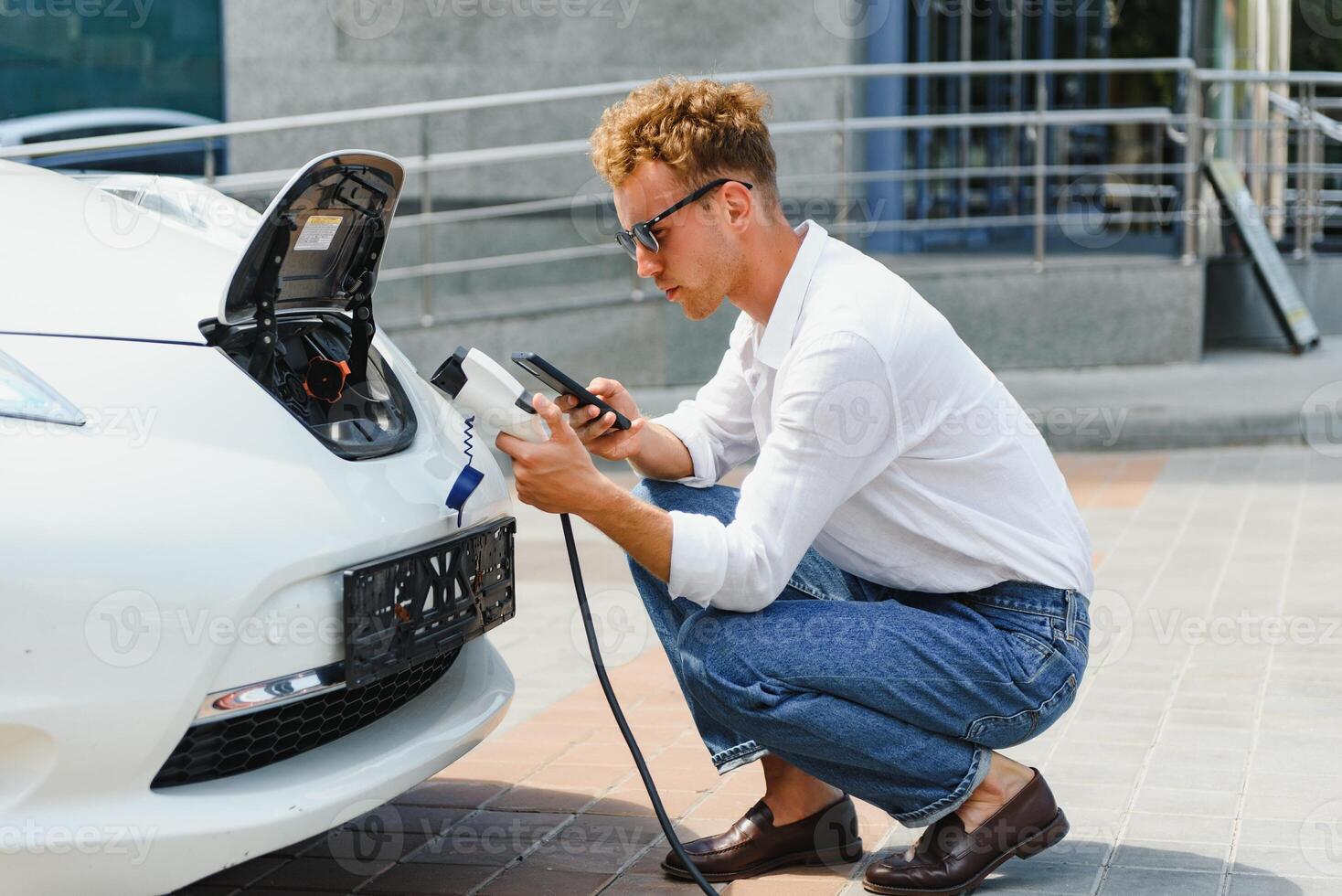 Casual man with smartphone near electric car waiting for the finish of the battery charging process photo
