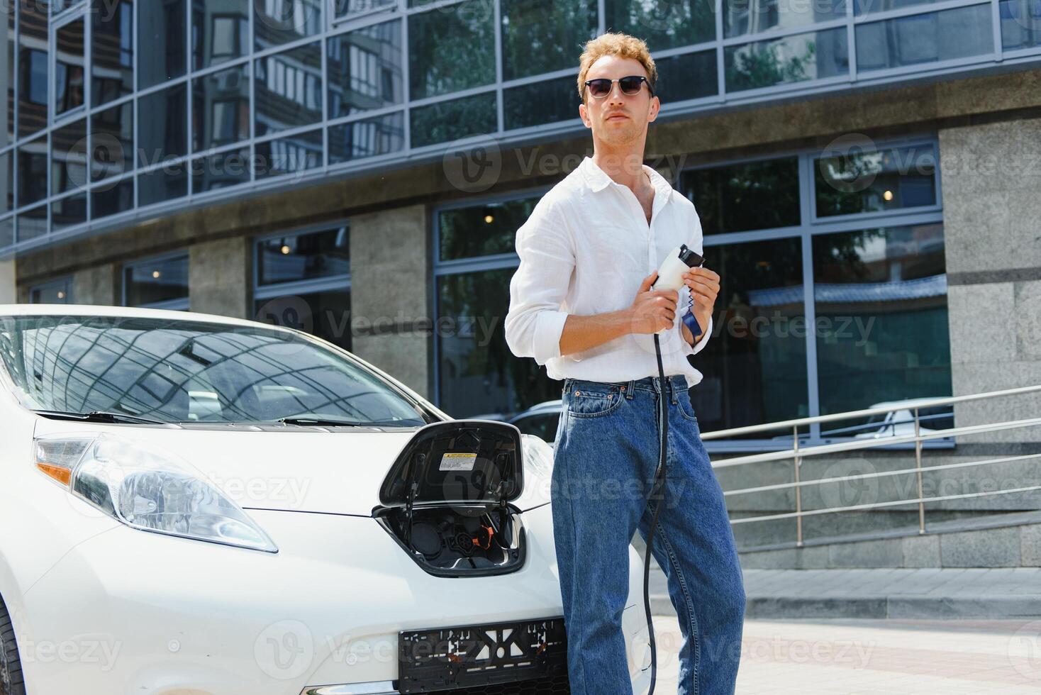 young handsome man holding charging cable at electric charging station point standing near his new car , looking satisfied. photo