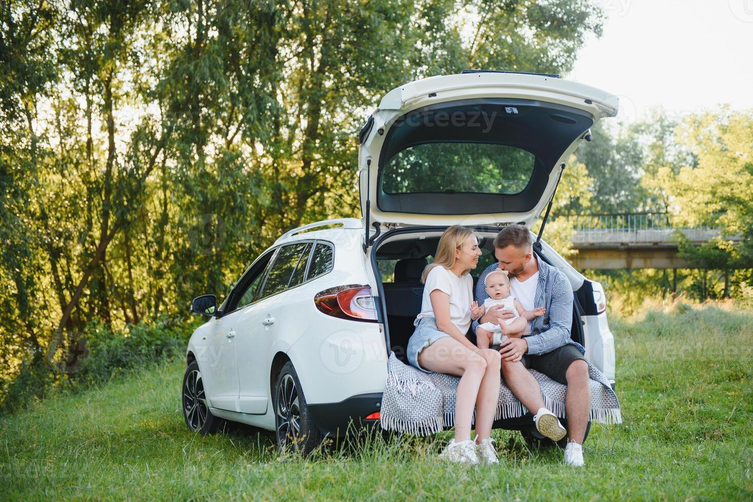 Pretty young married couple and their daughter are resting in the nature. The woman and girl are sitting on open car boot. The man is standing near them. They are smiling photo