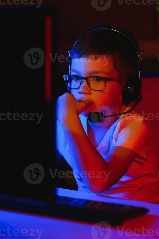 Child online broadcasts computer game, boy streams in headphones on rgb lighting background photo