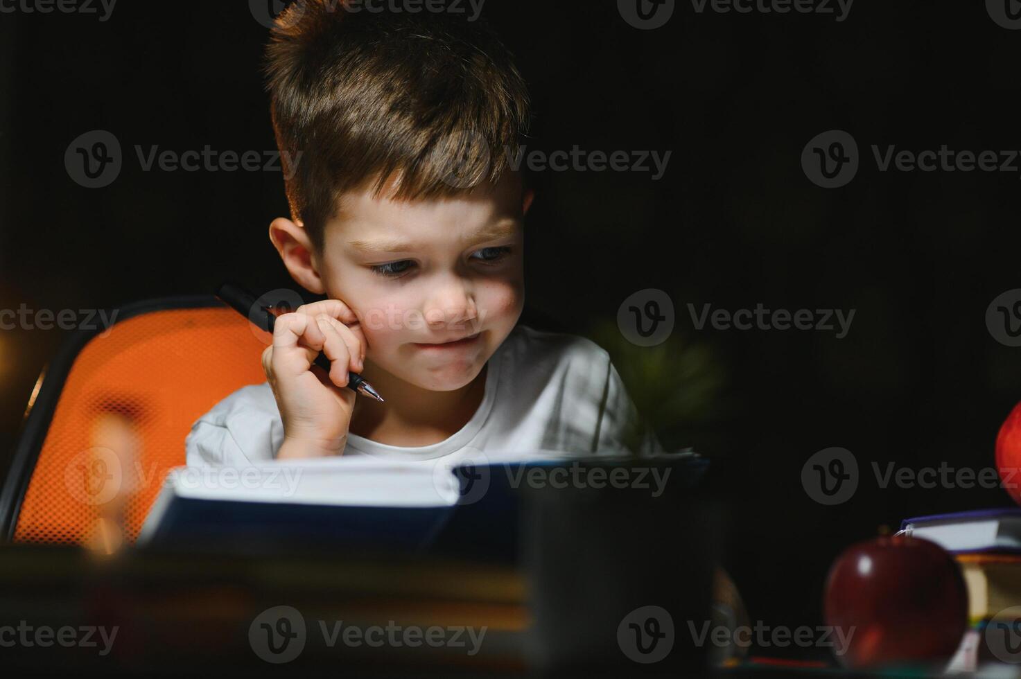 concentrated schoolboy reading book at table with books, plant, lamp, colour pencils, apple, and textbook photo