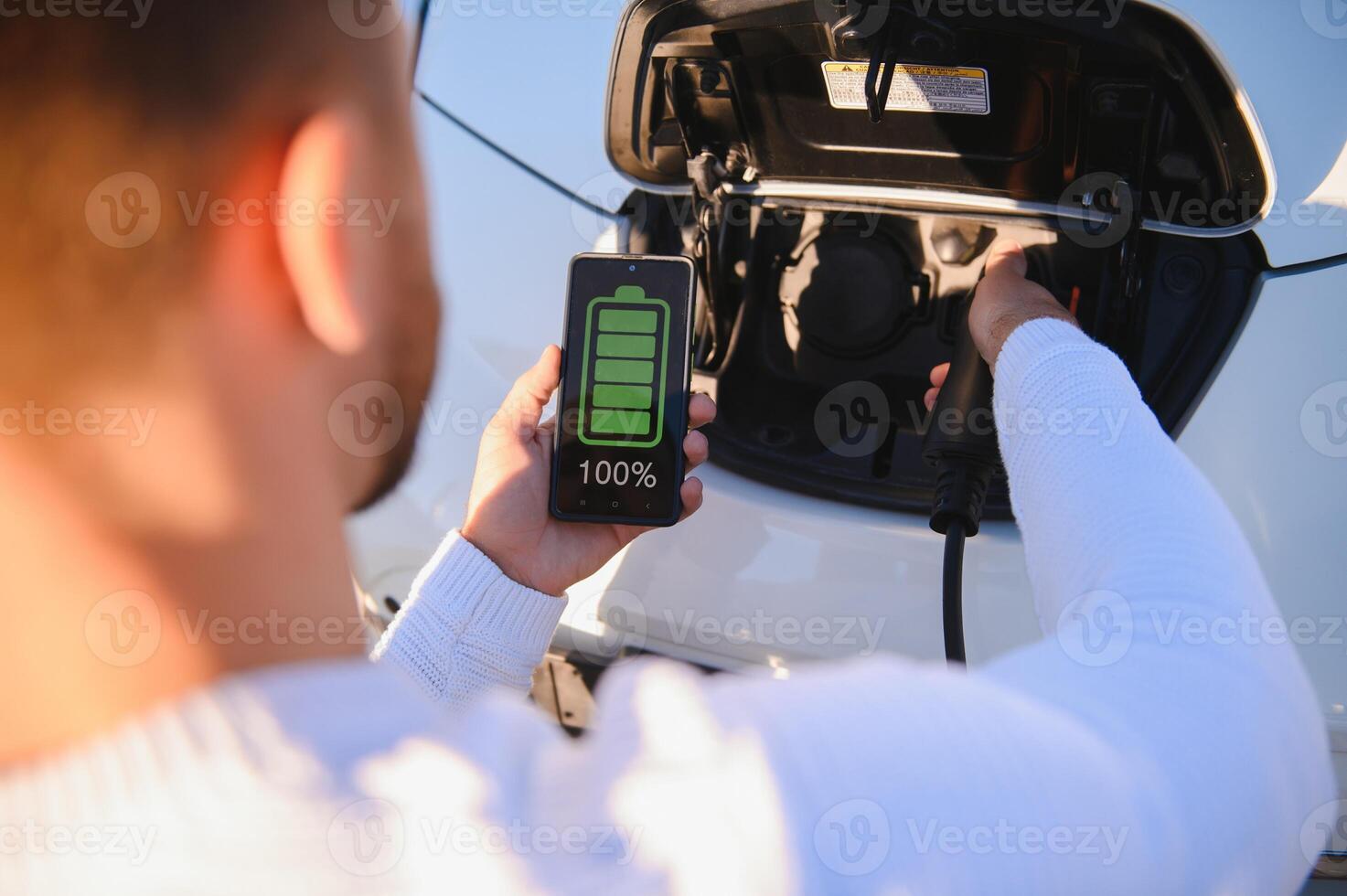 Man Charging Electric Vehicle With Cable Looking At App On Mobile Phone photo