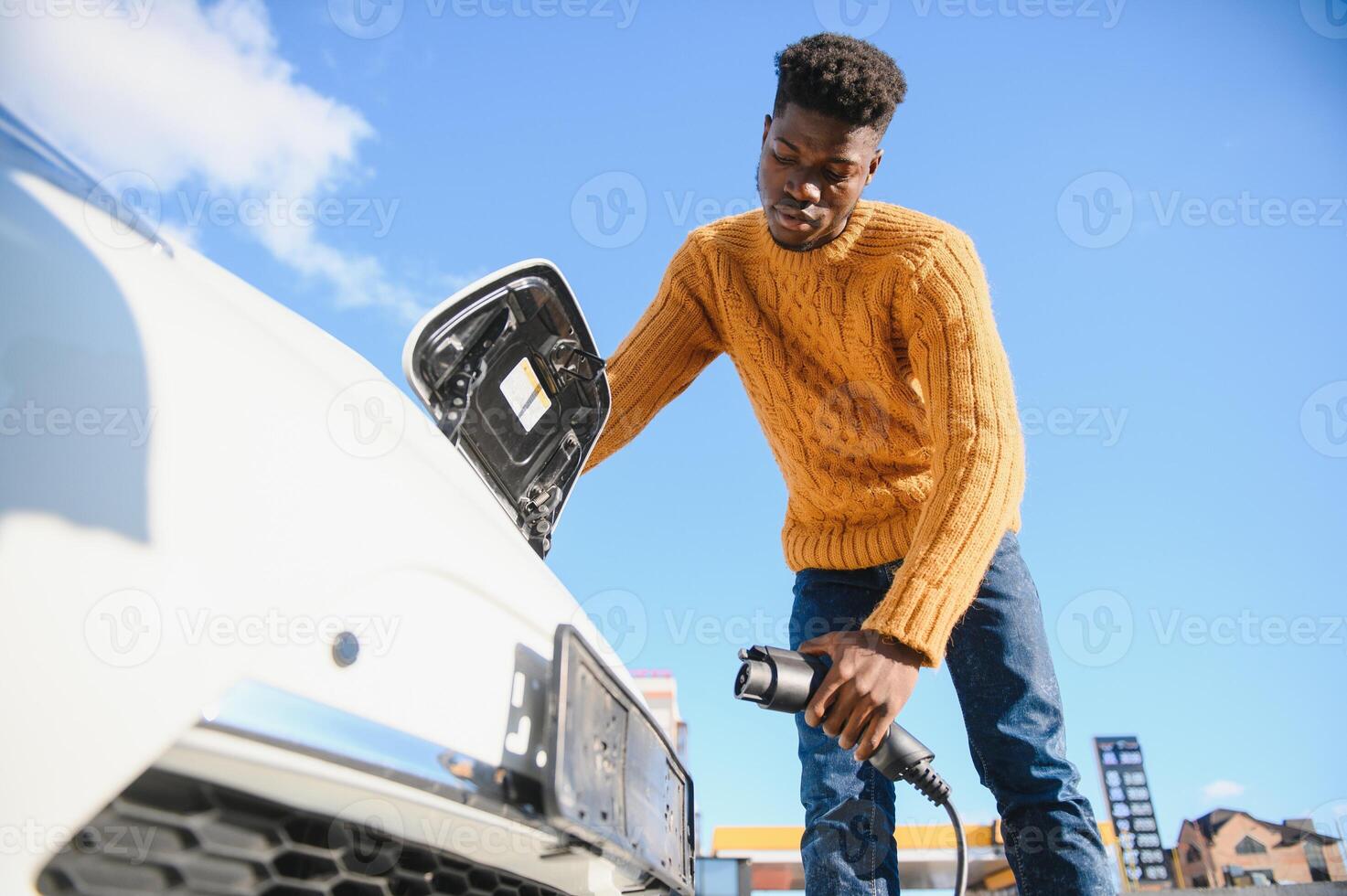 Close up of african american man connecting charging cable to electric car. Young male standing near his modern auto with leather suitcase in hand. photo