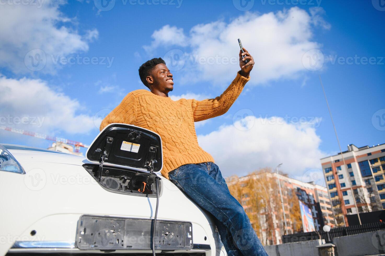 Portrait of young handsome black man in business suit, making selfie photo on his smartphone while leaning on his electric car, charging the battery at city power station