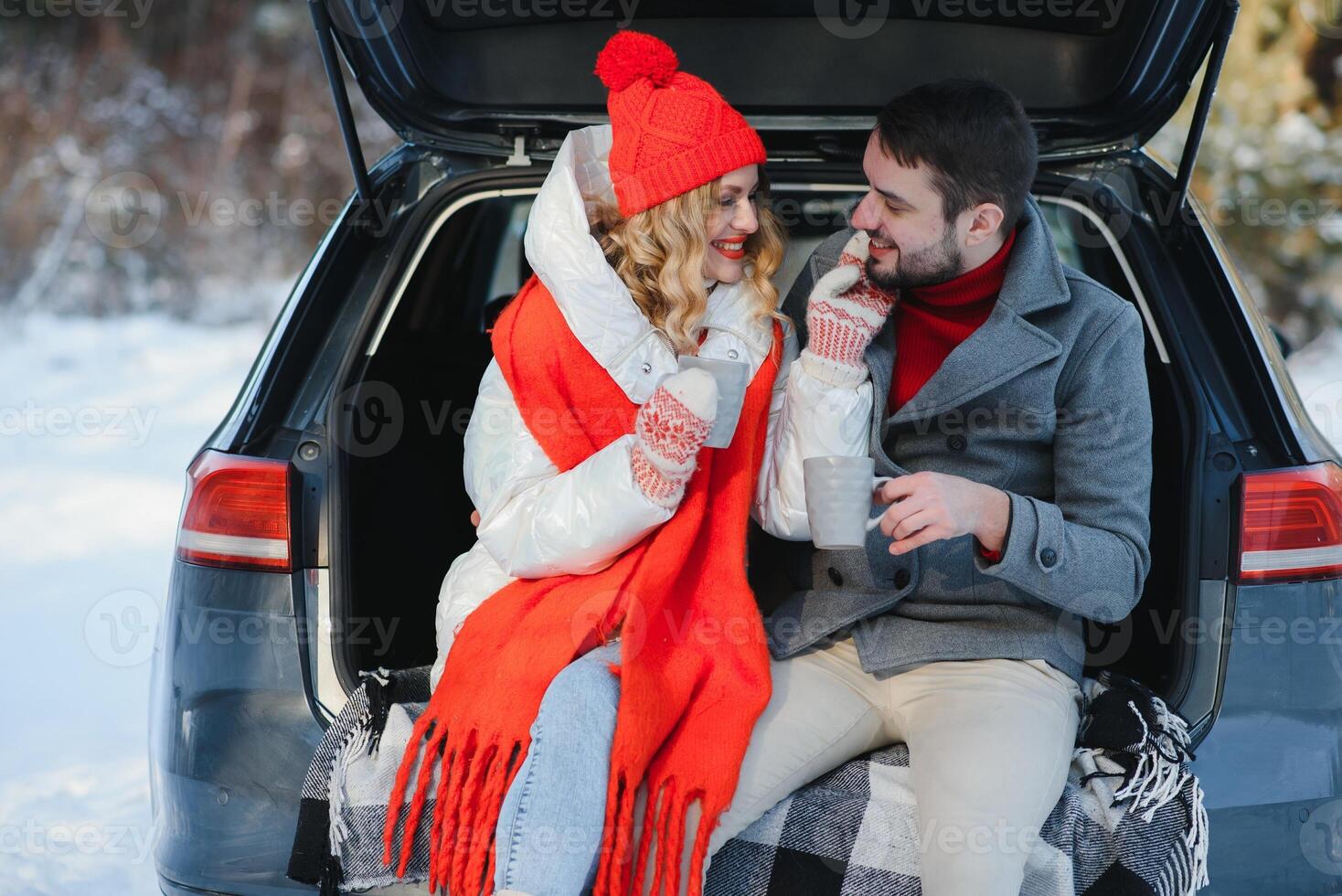 Happy Young couple is sitting in the back trunk of car in winter forest and drinking hot tea from thermos. Winter time photo