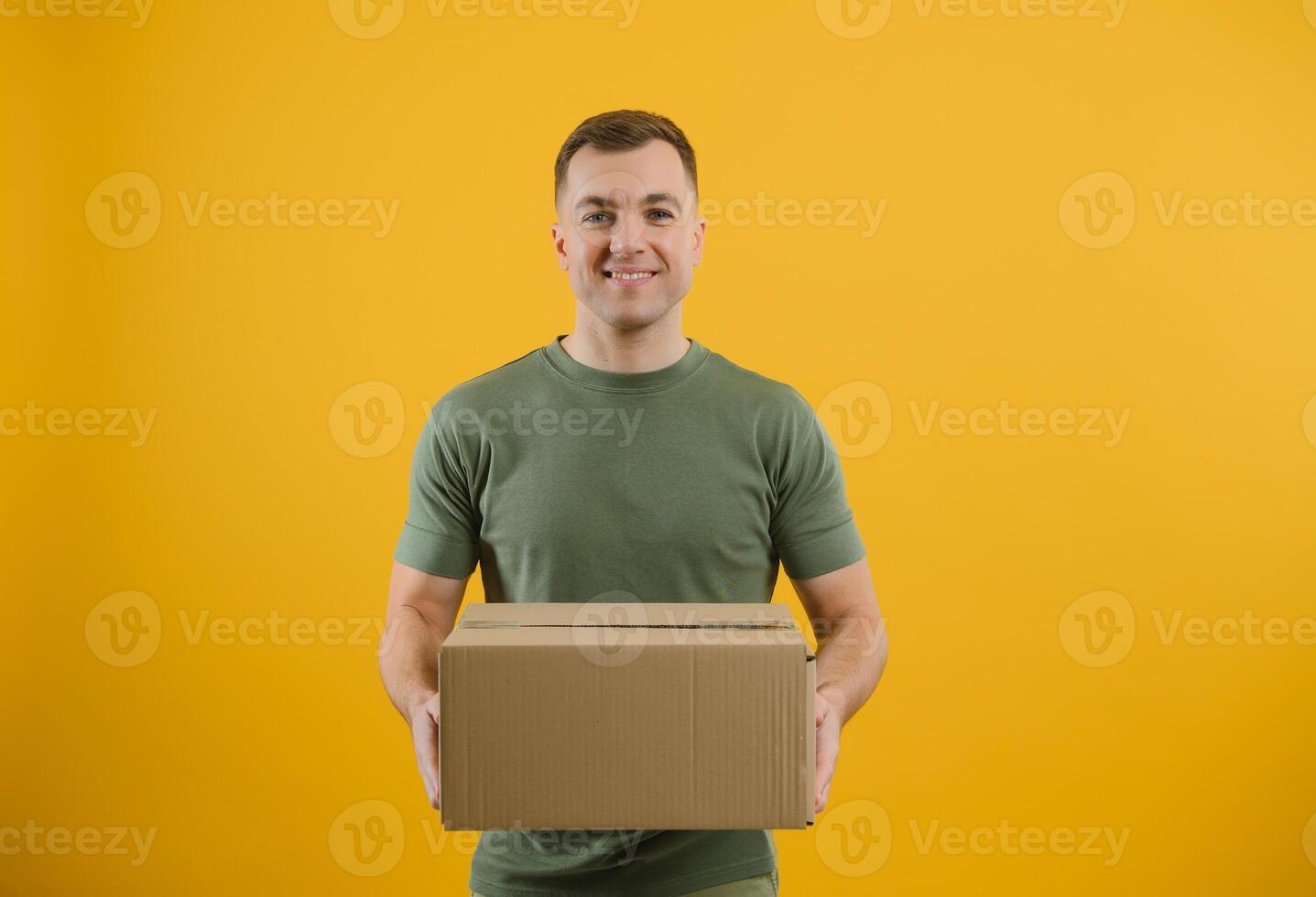 Delivery man in green uniform isolated on yellow background, studio portrait. Male employee working as courier dealer hold empty cardboard box. Service concept. Mock up copy space photo