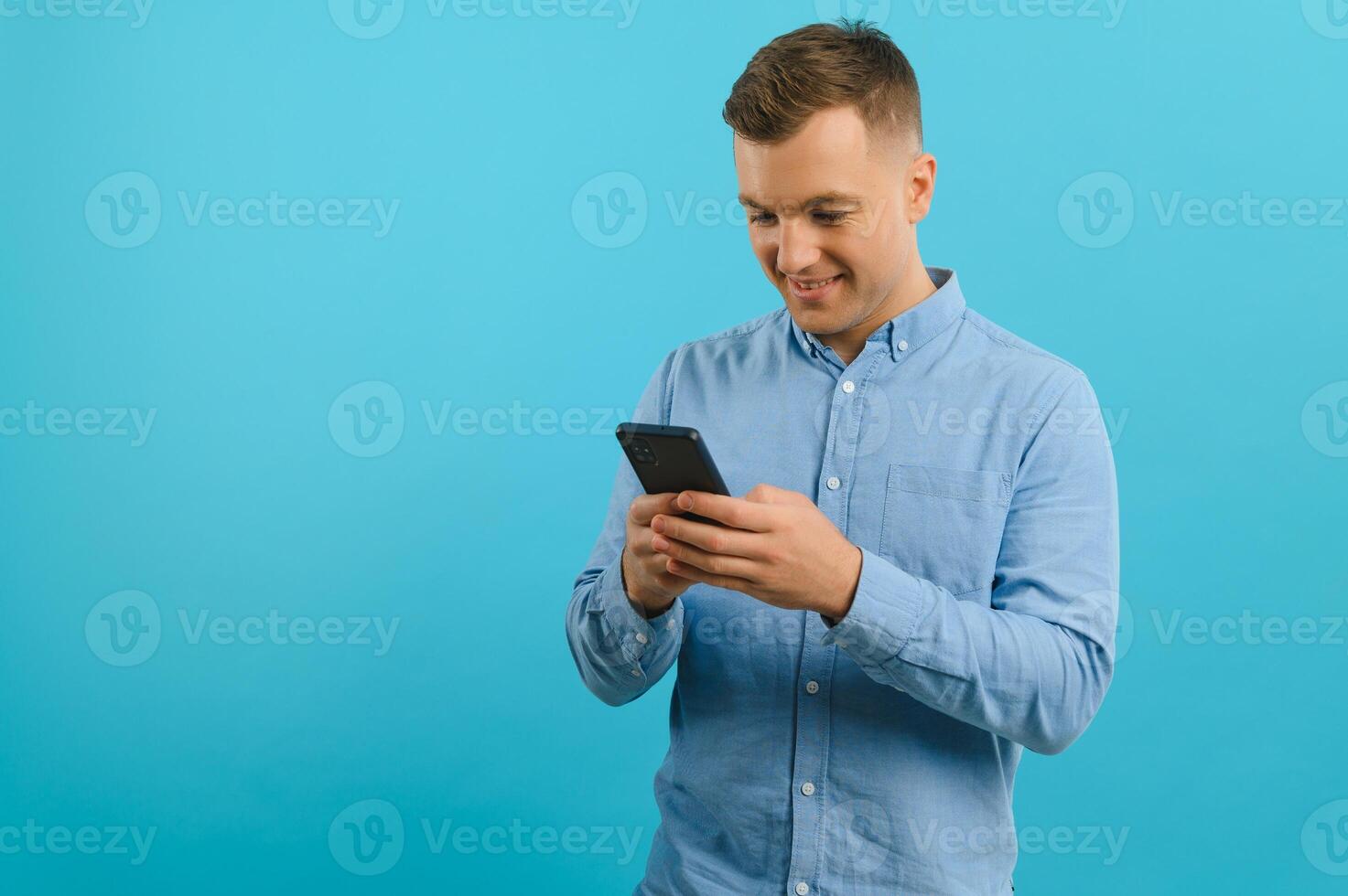 Photo of happy handsome man holding mobile phone in hand, texting. Young guy with big toothy smile posing on blue background.