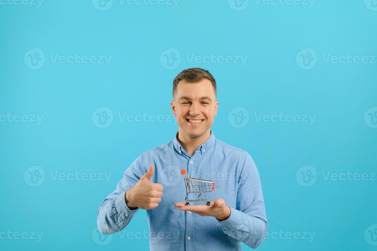 Young smiling man holding a mini shopping cart on a blue background. Sales concept. Place for text. photo