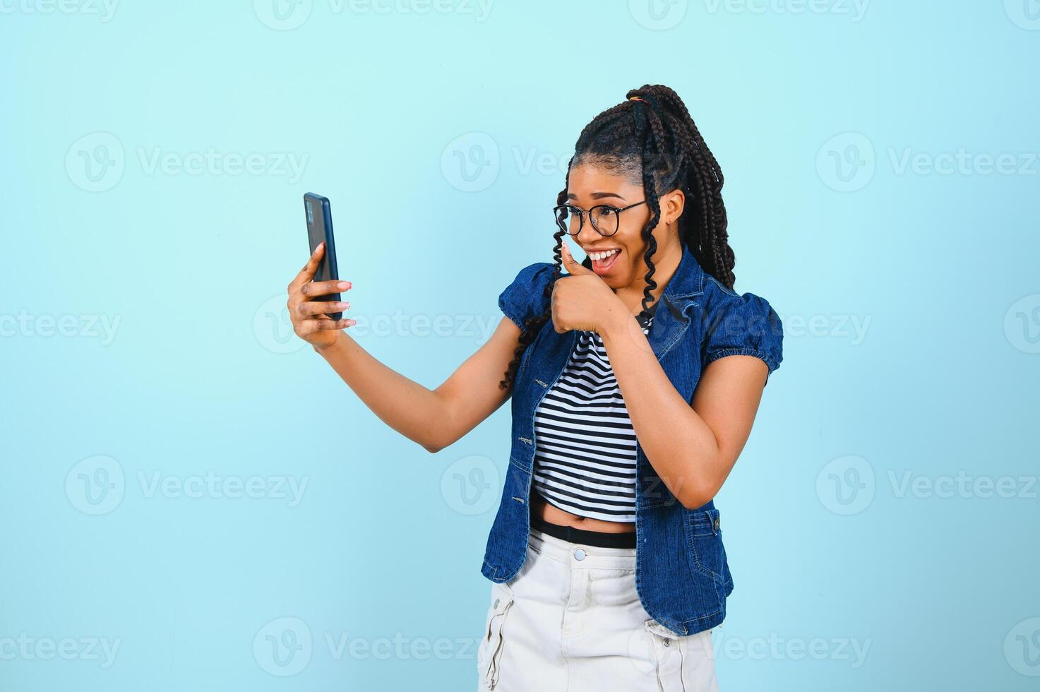 Happy cheerful Afro American woman looks at screen of smart phone enjoys online chatting types text message surfs social networks dressed casually poses against blue background. Technology concept. photo