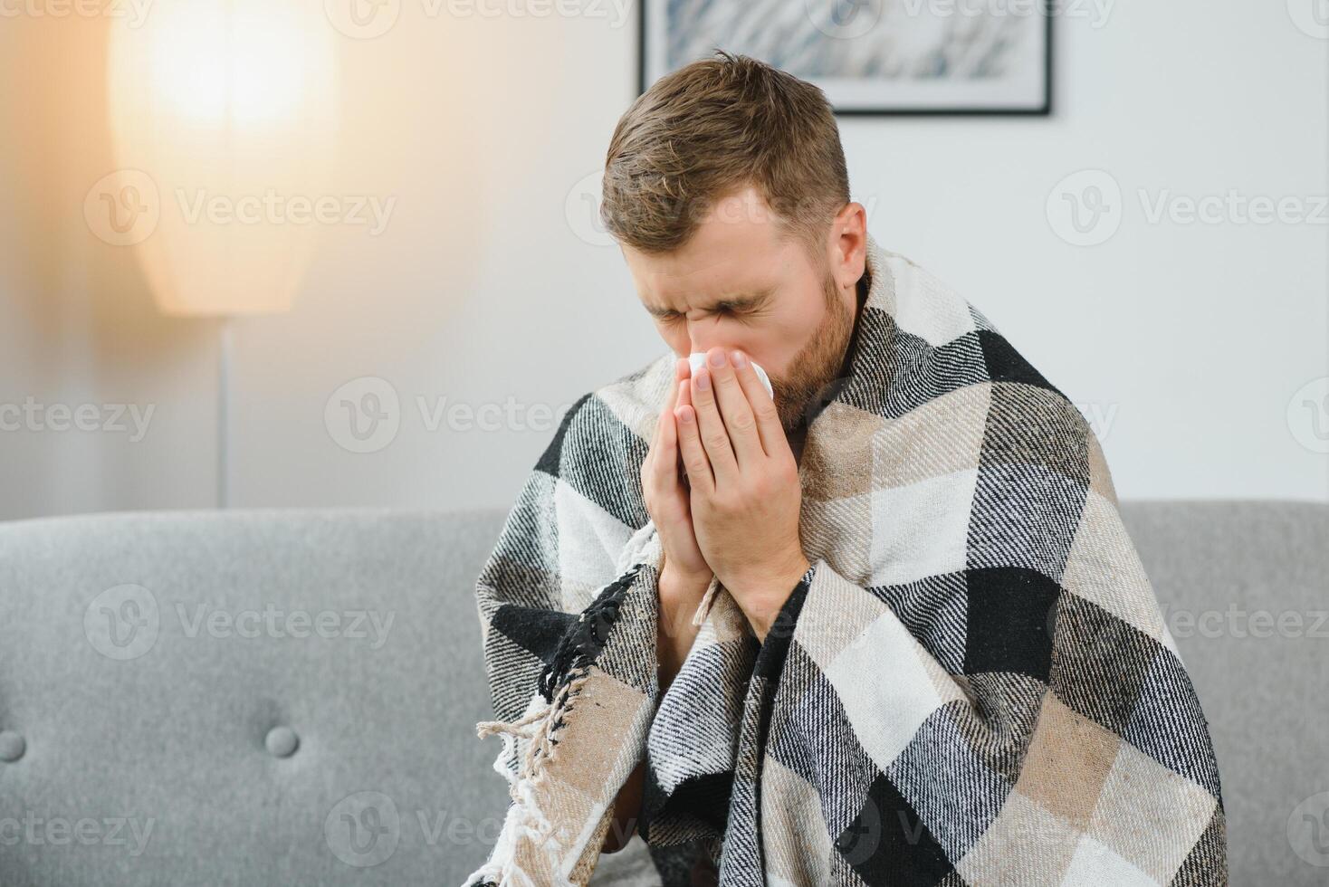 Sick adult man who has bad cold or seasonal flu virus is fighting illness at home. Unhappy middle aged man is sitting on sofa wrapped in warm scarf and plaid, holding paper tissue and sneezing. photo