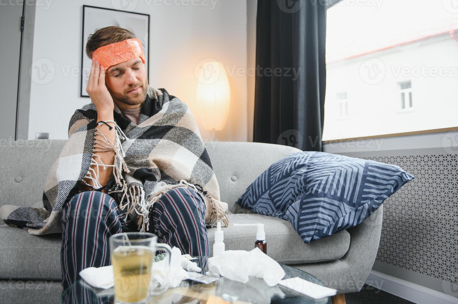 Sick bearded man who has bad cold or seasonal flu sitting on couch at home. Guy with fever wearing warm plaid shivering with worried face expression. photo