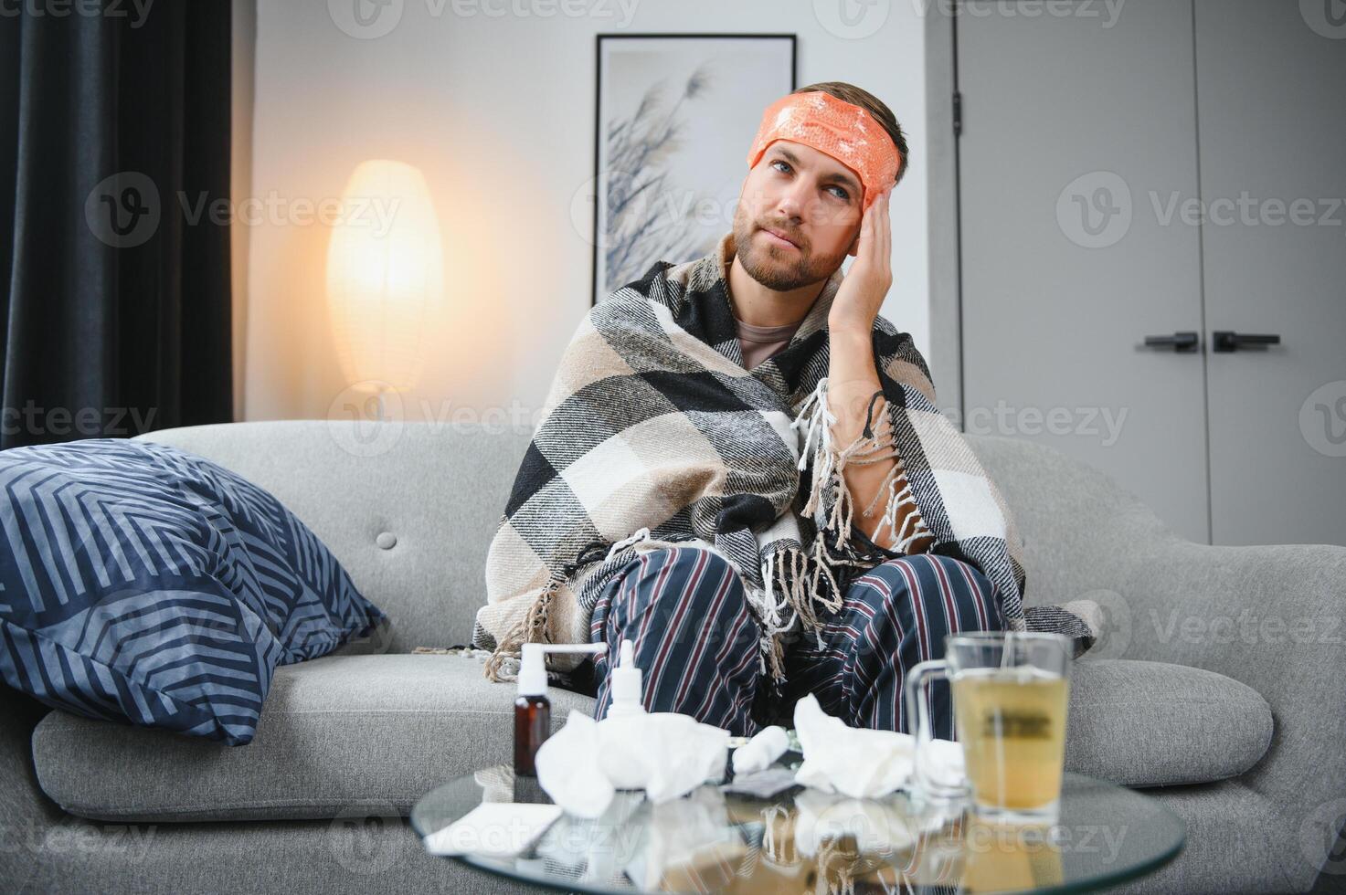 Sick bearded man who has bad cold or seasonal flu sitting on couch at home. Guy with fever wearing warm plaid shivering with worried face expression. photo