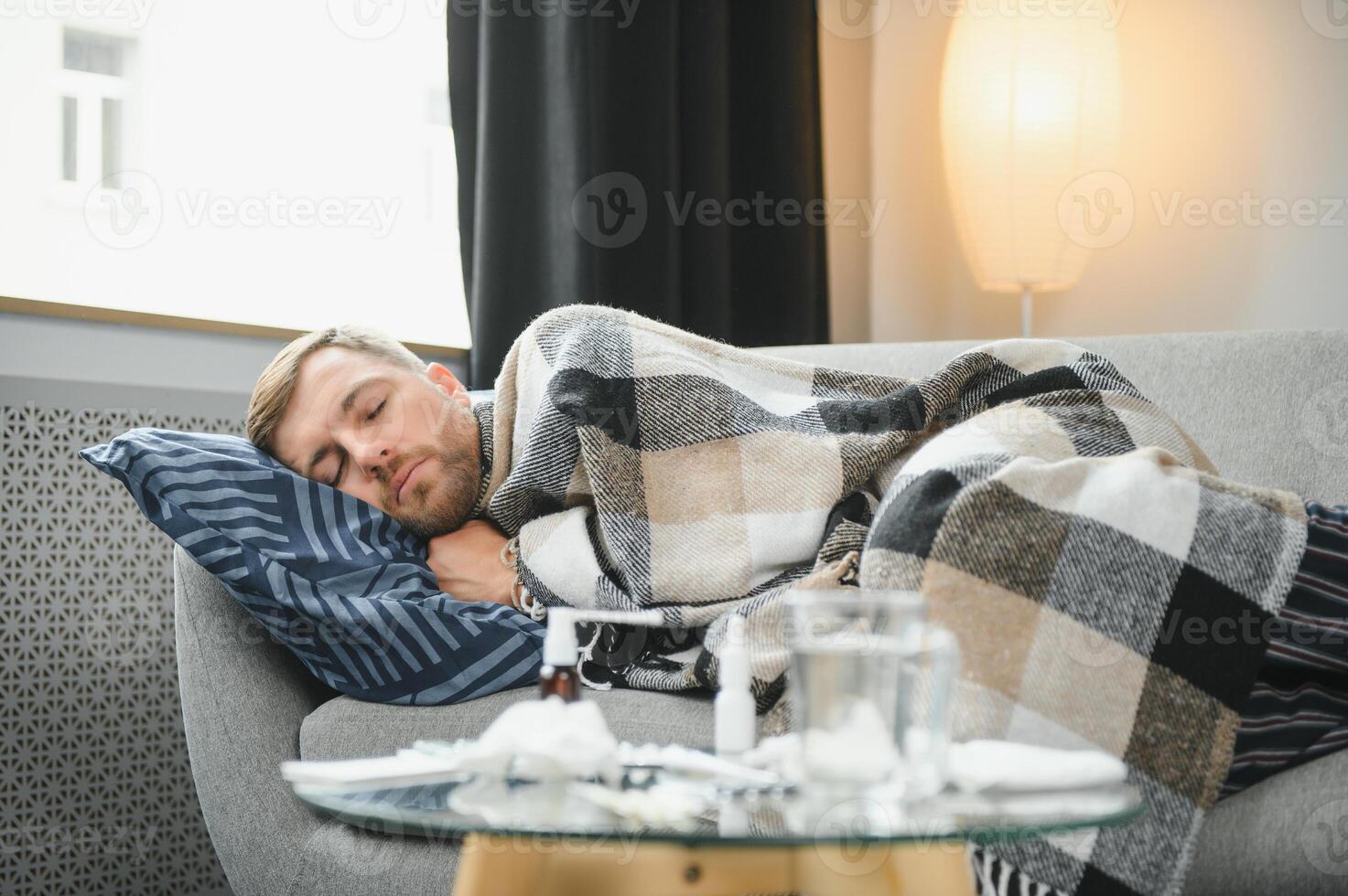 Sick bearded man who has bad cold or seasonal flu sitting on couch at home. Guy with fever wearing warm plaid shivering with worried face expression. photo