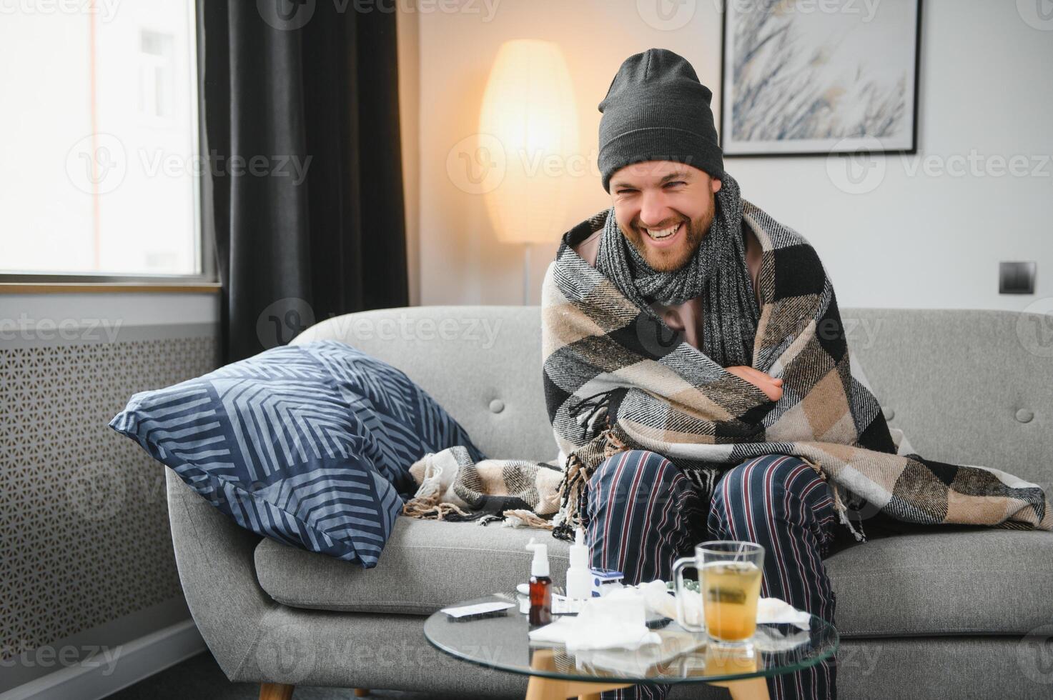 Sick bearded man who has bad cold or seasonal flu sitting on couch at home. Guy with fever wearing warm plaid shivering with worried face expression. photo