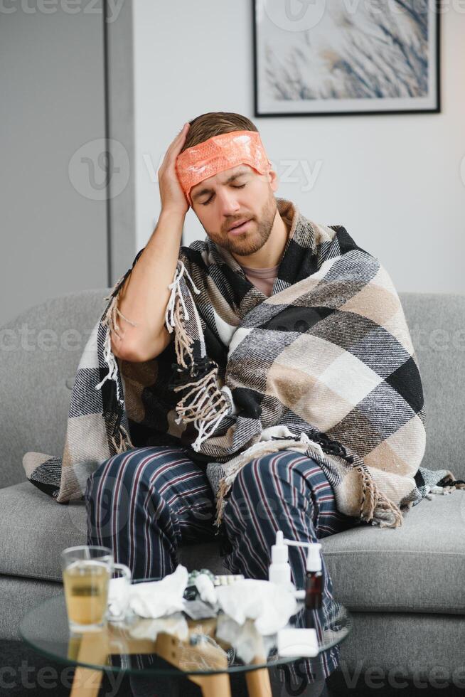 Sick adult man who has bad cold or seasonal flu virus is fighting illness at home. Unhappy middle aged man is sitting on sofa wrapped in warm scarf and plaid, holding paper tissue and sneezing. photo
